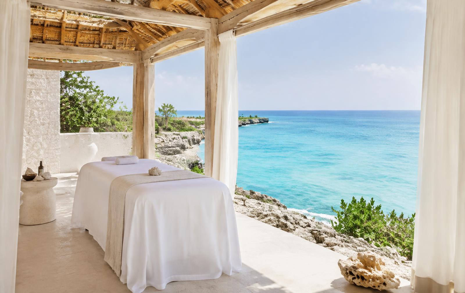 Open-air timber spa pavilion with white linen above the turquoise ocean